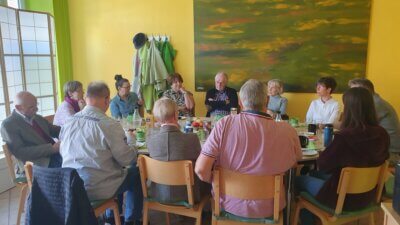 Ca ein dutzend Personen sitzen um einen runden Tisch, ein gelbe Wand und ein abstraktes grünes Gemälde im Hintergrund